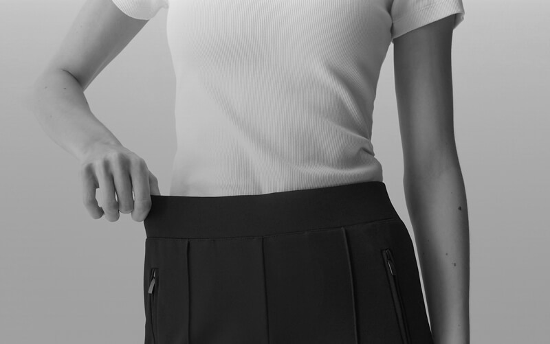 Black and white close-up shot of a person wearing a white ribbed t-shirt and a black OPUS PANTS. The frame captures the torso from waist to chest, with the person's hand resting on their waist. Due to the Size Bridge the pants fits very well.